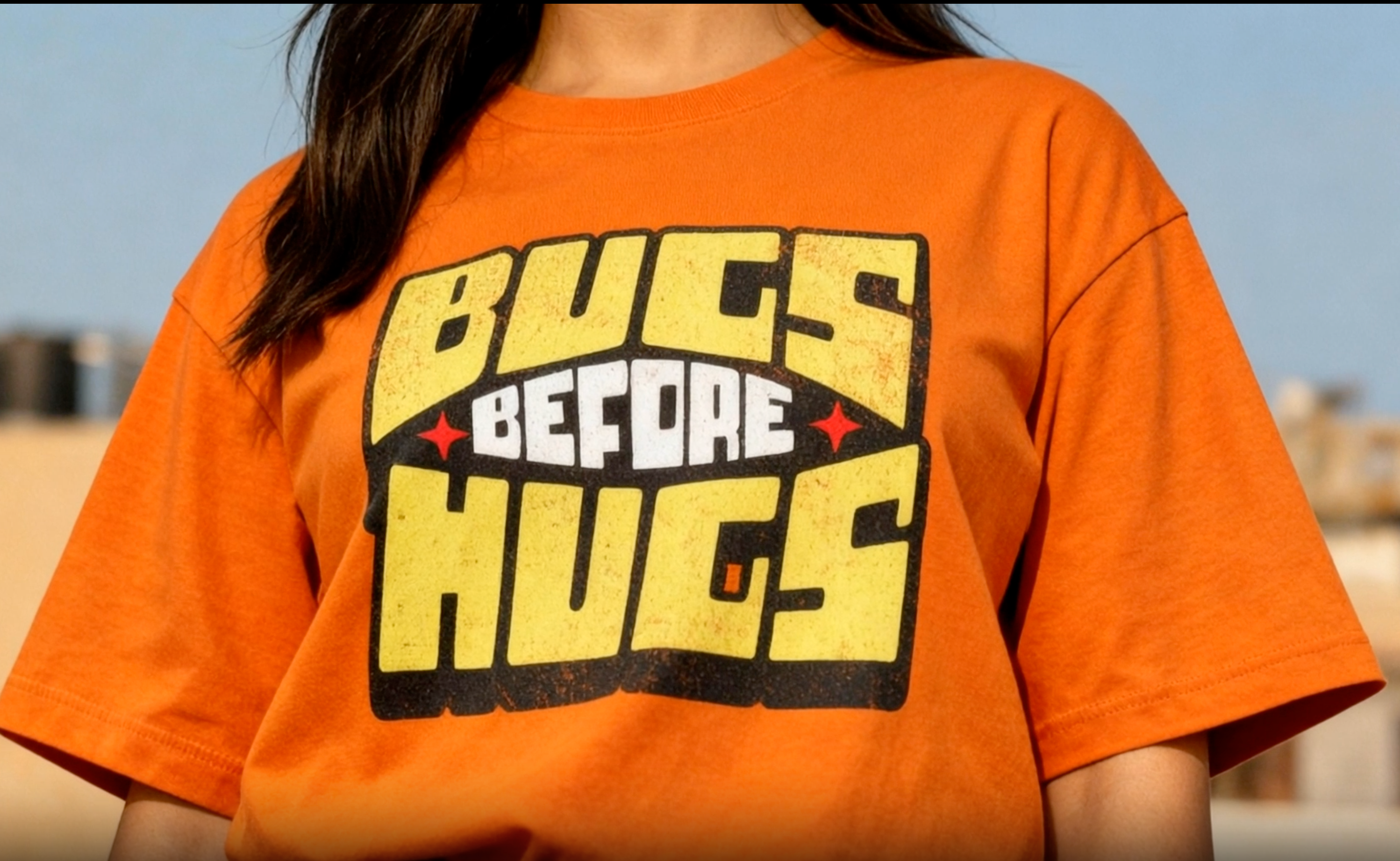 Person wearing an orange t-shirt with 'Bugs Before Hugs' text.