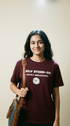Person wearing a maroon t-shirt with text and a clock graphic, holding a bag against a plain background
